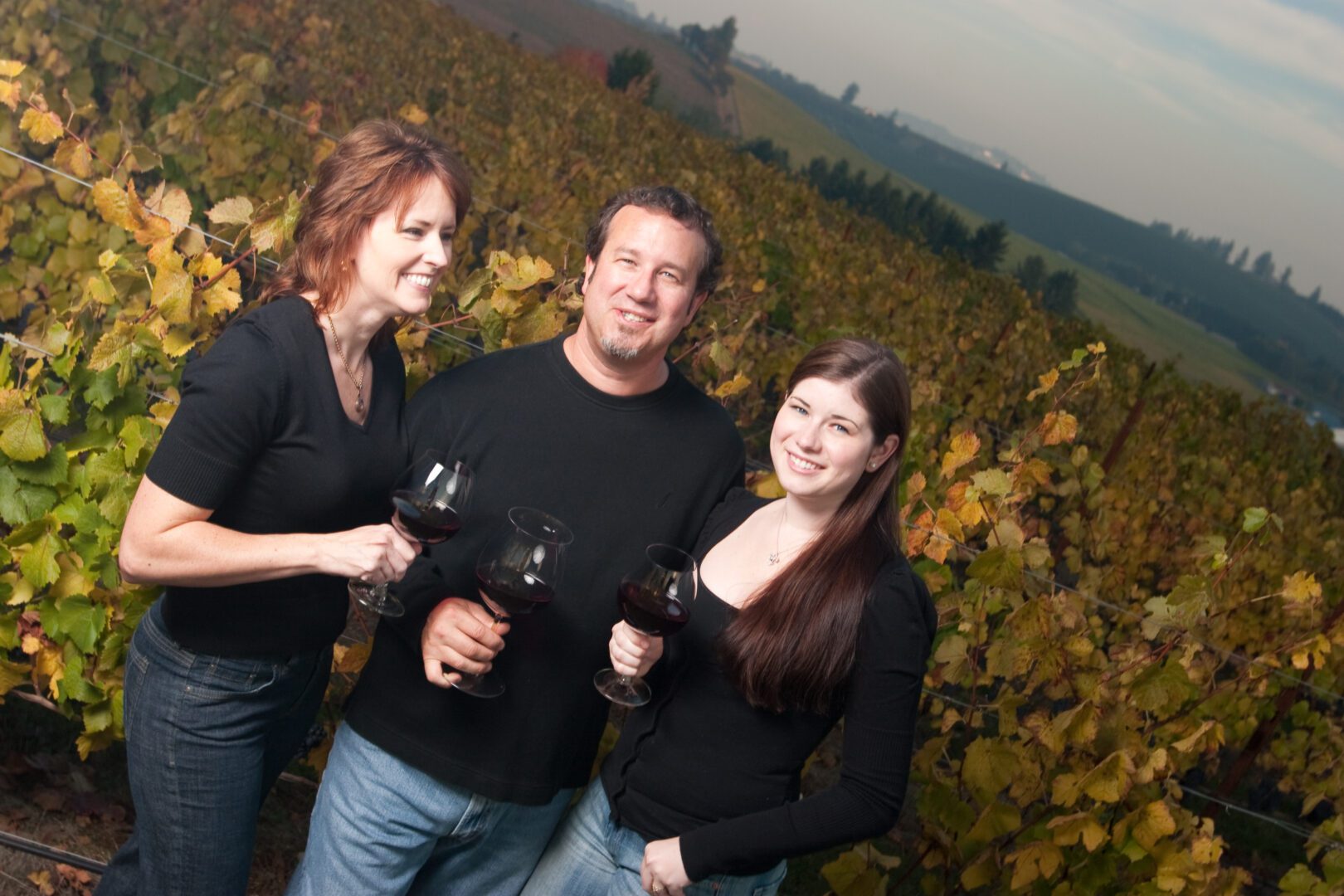 Three people holding wine glasses in a vineyard.