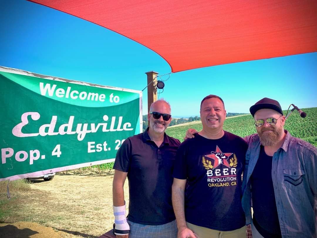 Three men standing under a red umbrella in front of a sign.